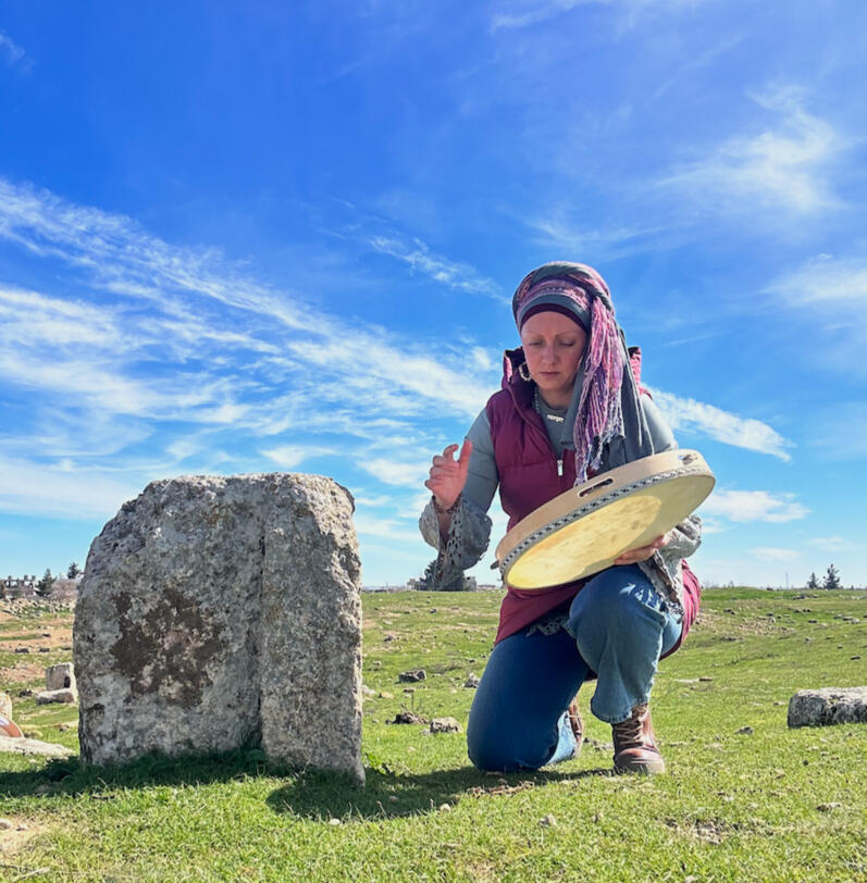 Lisa Moriah in ritual practice on the ground at Harran A woman kneeling with an Anatolian tambourine next to a broken foundation stone in the archaeological field at Harran, February 2024